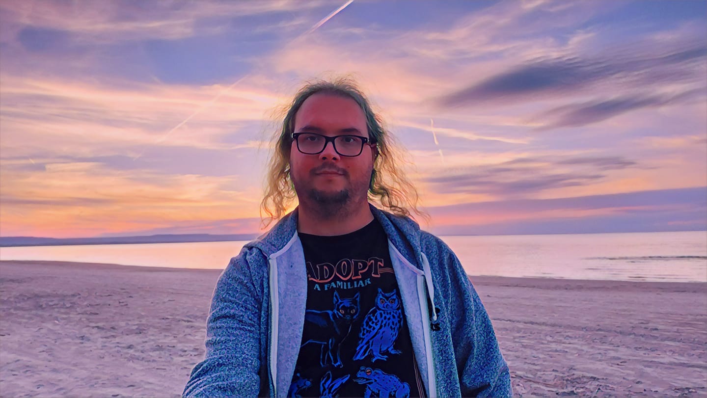 A man with green hair wearing a swetter sits on the soft white sand, in the background Lake Huron and the Blue Mountains, the sky is bright with the colours of sunset